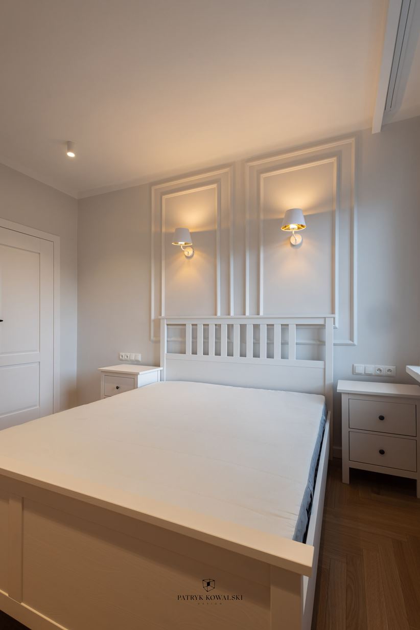 Biała sypialnia ze sztukateriami i kinkietami nad drewnianym łóżkiem